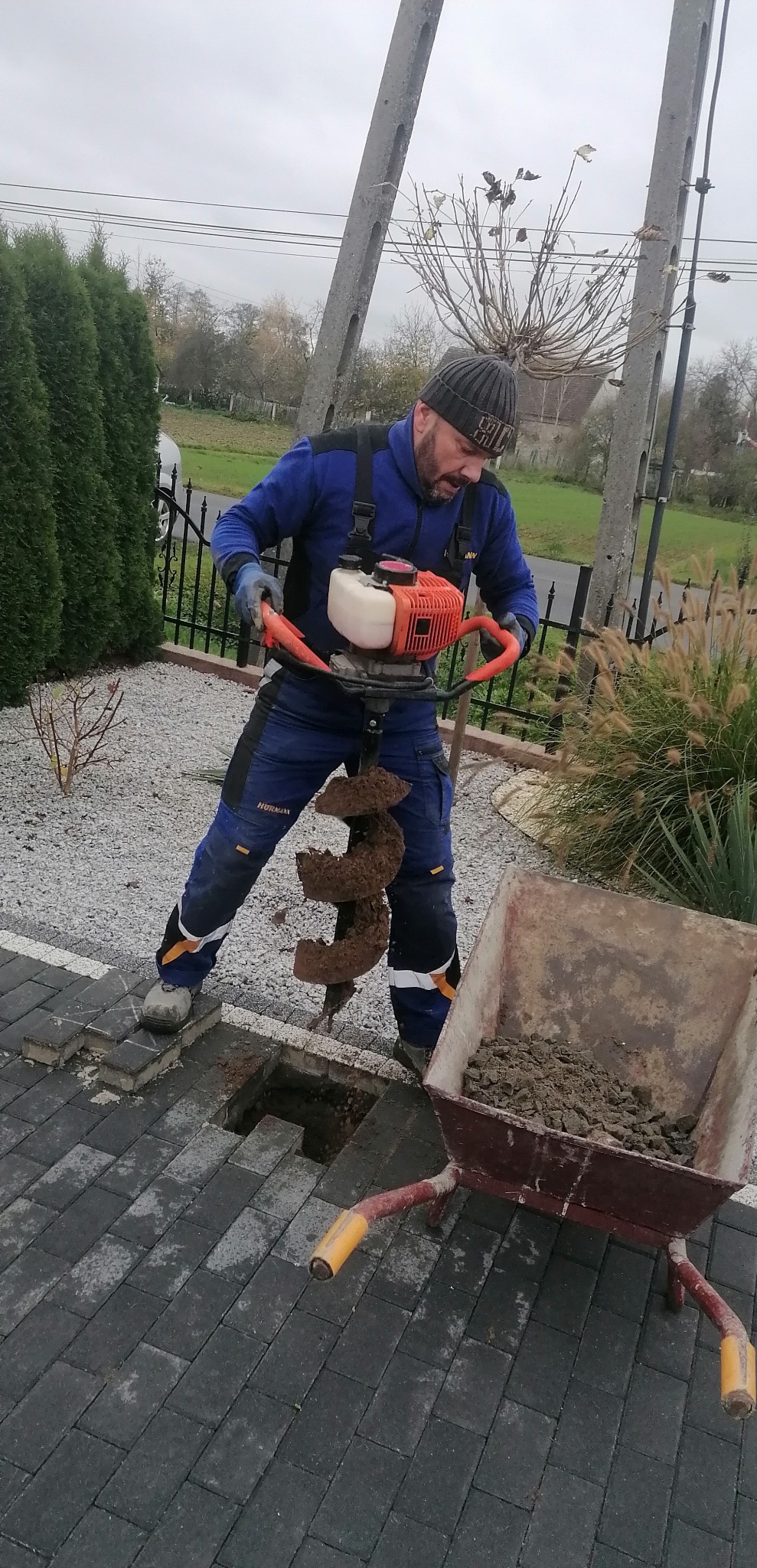 Mężczyzna wierci otwór w ziemi wiertnicą spalinową na kostce brukowej. Obok taczka z ziemią. W tle ogrodzenie i zieleń. Praca w ogrodzie.