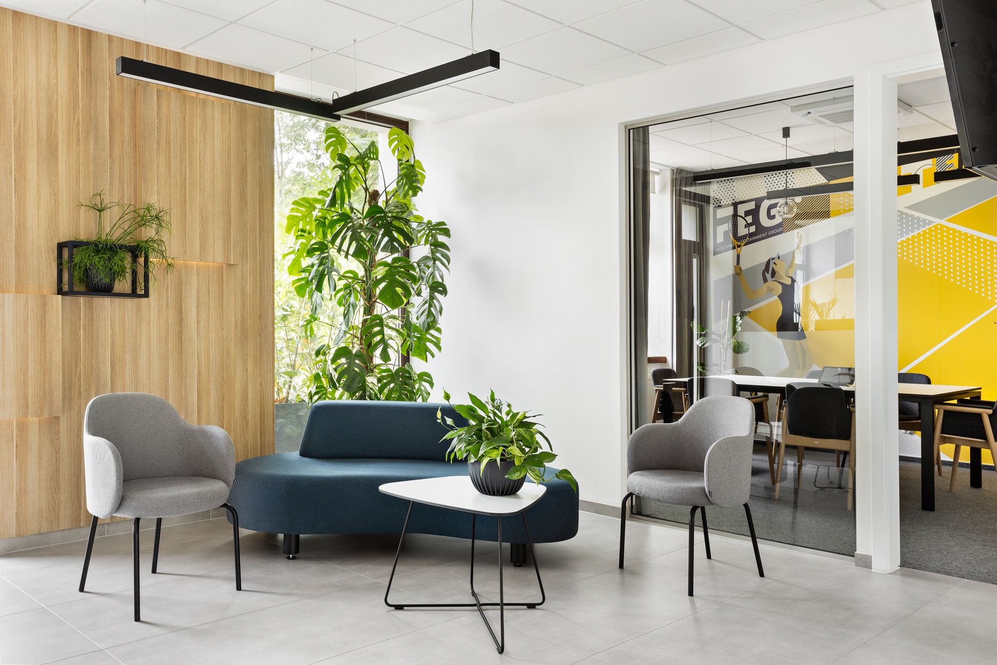 Stylowa aranżacja biura z sofą, fotelami i roślinami. Widoczna przeszklona sala konferencyjna z żółto-szarą grafiką na ścianie. Nowoczesne oświetlenie.