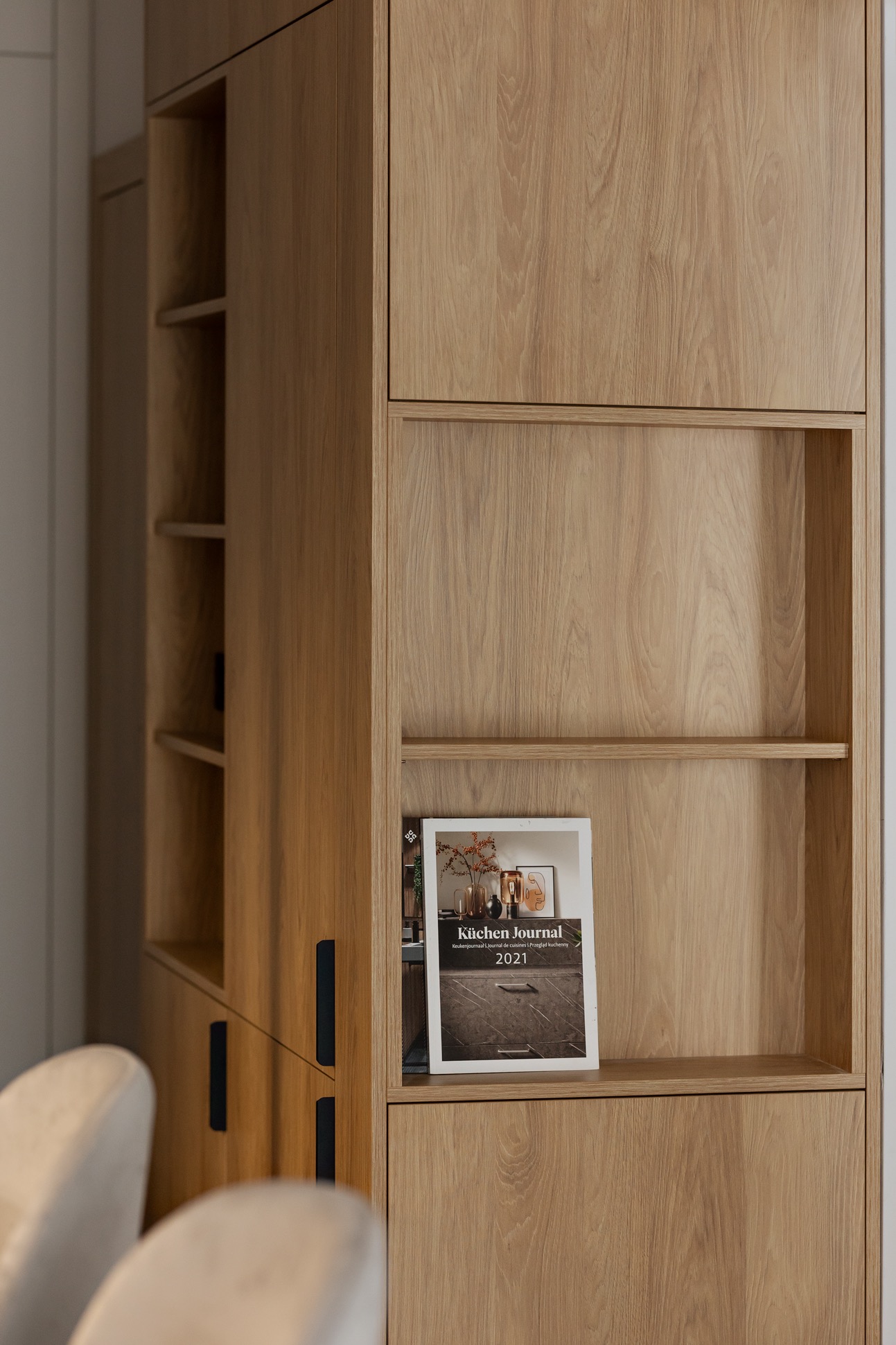 Jasna, drewniana szafa z półkami i czarnymi uchwytami, z magazynem 'Küchen Journal' na półce. Minimalistyczny design, ciepłe oświetlenie, widoczne krzesła w tle.