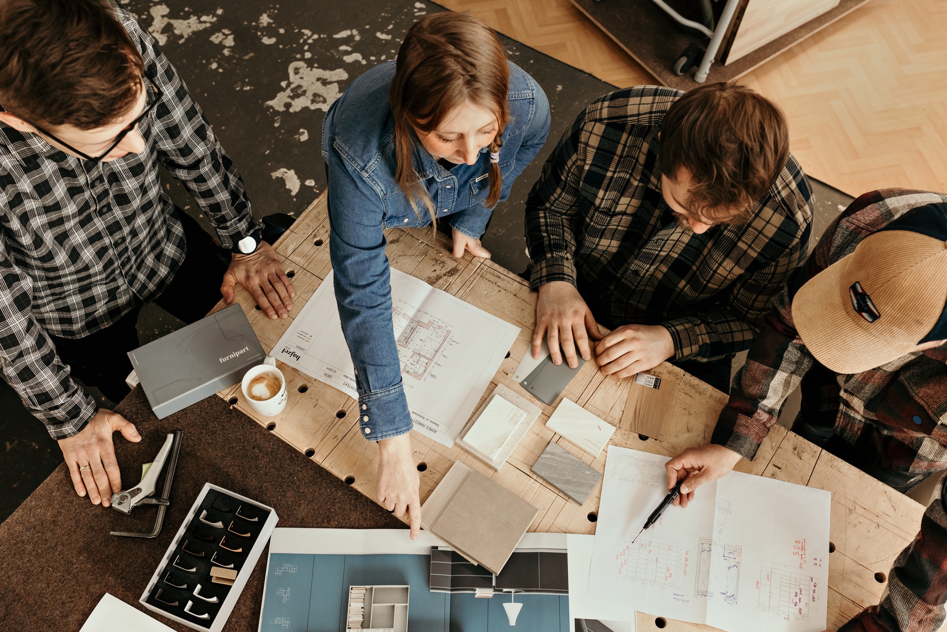 Zespół projektowy analizuje plan i próbki materiałów na drewnianym stole. Widok z góry ukazuje detale projektu i wybór wykończeń, w tym uchwyty meblowe i paletę kolorów.