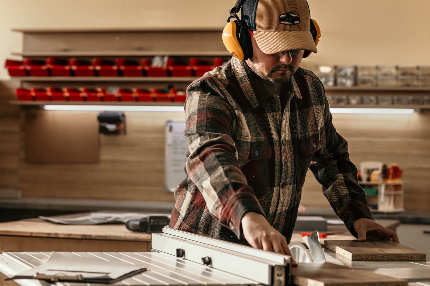Stolarz w słuchawkach tnący płytę na pile stołowej w warsztacie. Widoczne pojemniki z narzędziami i oświetlenie LED. Ujęcie z góry, skupione na pracy.