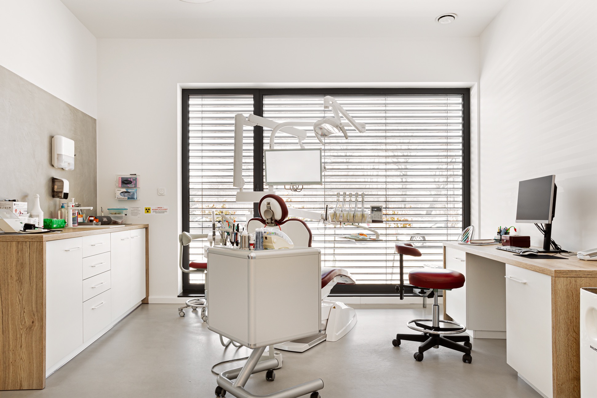 Nowoczesny gabinet stomatologiczny z białymi meblami, fotelem dentystycznym i dużym oknem z żaluzjami, zapewniającym naturalne światło.