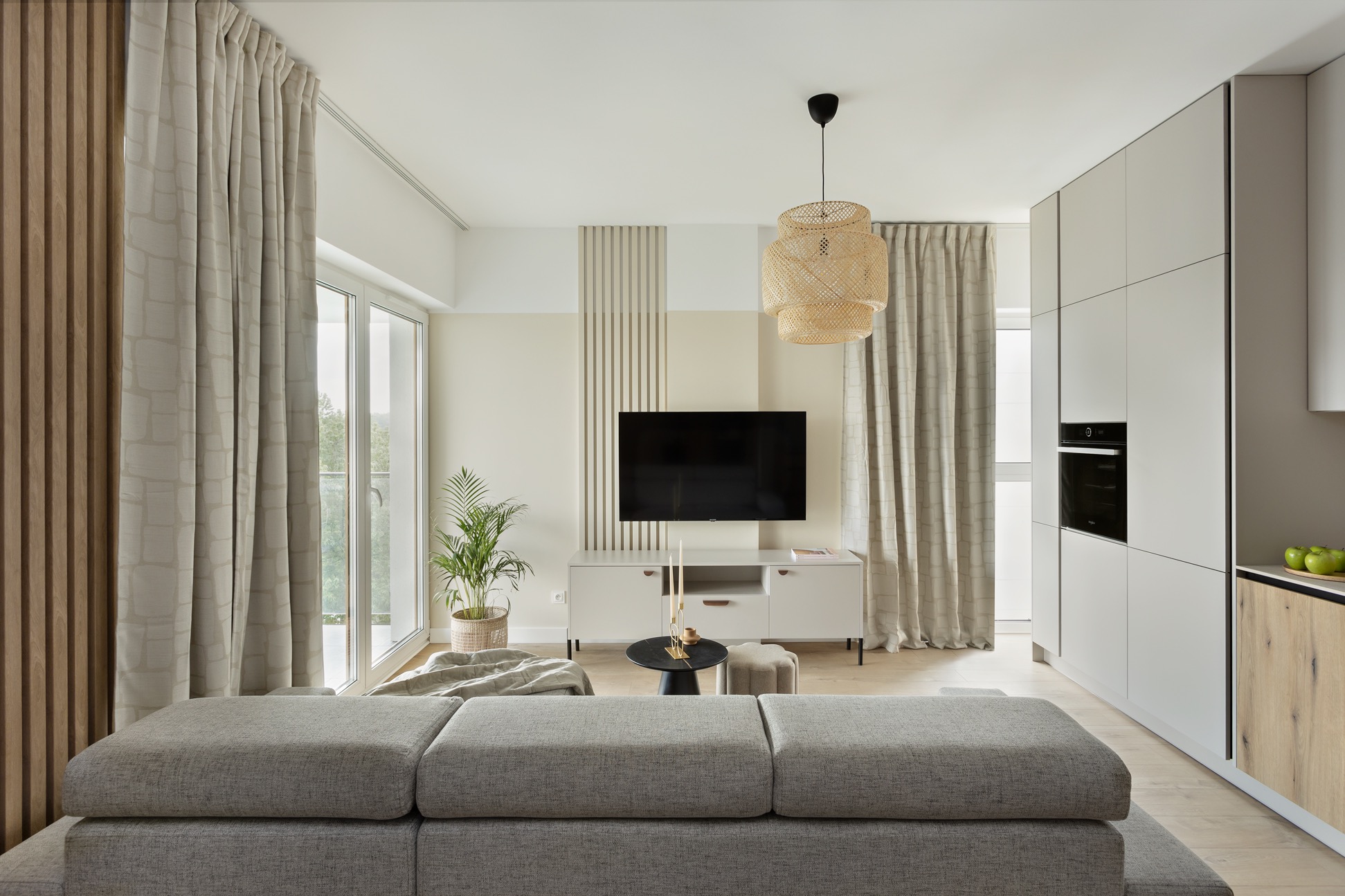 Jasne wnętrze salonu z aneksem kuchennym: szara sofa, białe szafki kuchenne z drewnianym akcentem, rattanowa lampa, okno z zasłonami i dekoracyjne panele ścienne.