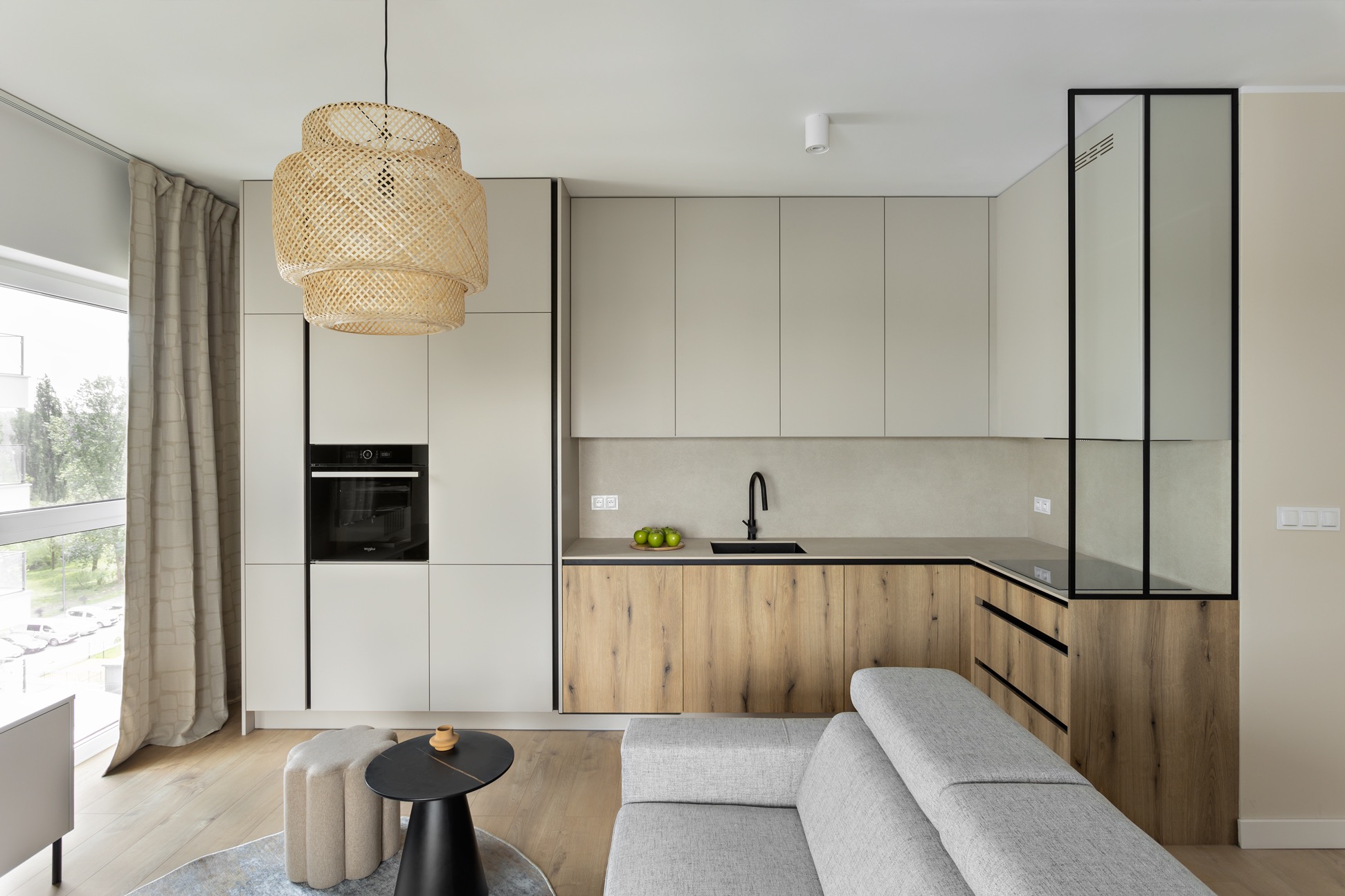 Jasna kuchnia z drewnianymi frontami szafek dolnych i beżowymi górnymi, oświetlona lampą z naturalnego materiału. Widoczny fragment sofy i stolika kawowego.