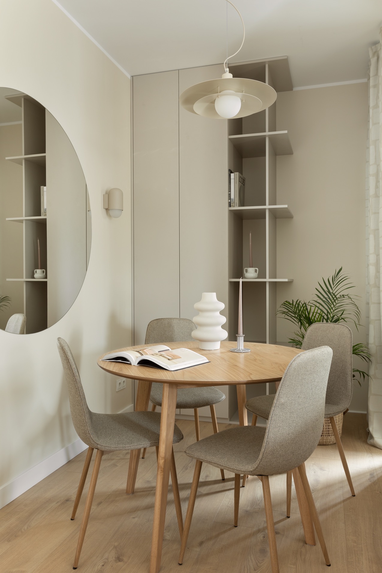 Jasne wnętrze jadalni: okrągły stół z książką, cztery krzesła, minimalistyczna lampa i regał w zabudowie. Lustro odbija fragment wnętrza. Neutralna kolorystyka.