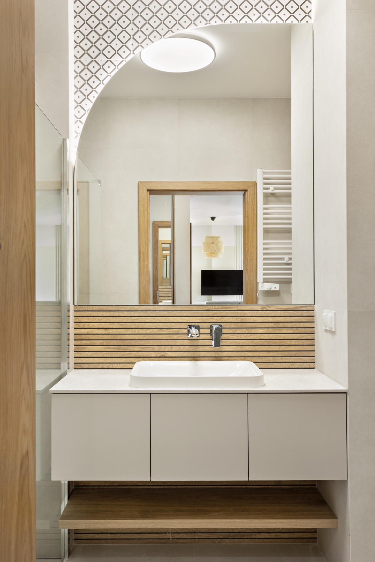 Nowoczesna łazienka z szafką pod umywalką w odcieniach beżu i drewna. Lustro z podświetlanym wzorem. Widok na wnętrze przez lustrzane odbicie.