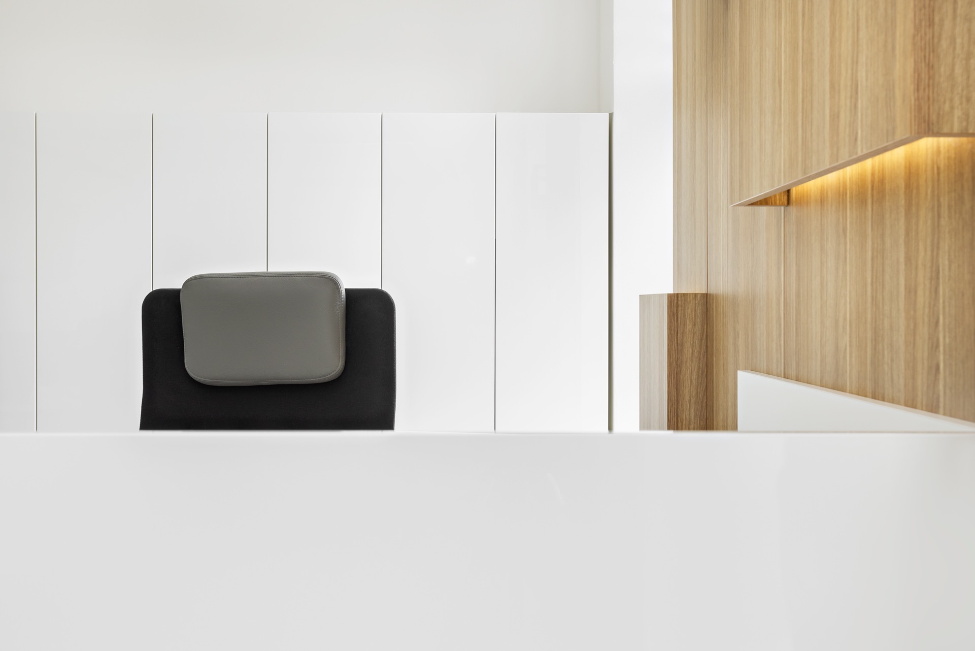 Minimalistyczne biuro z białymi szafkami, drewnianą ścianą z oświetleniem i czarnym krzesłem z szarą poduszką. Nowoczesny design, czyste linie, jasne wnętrze.