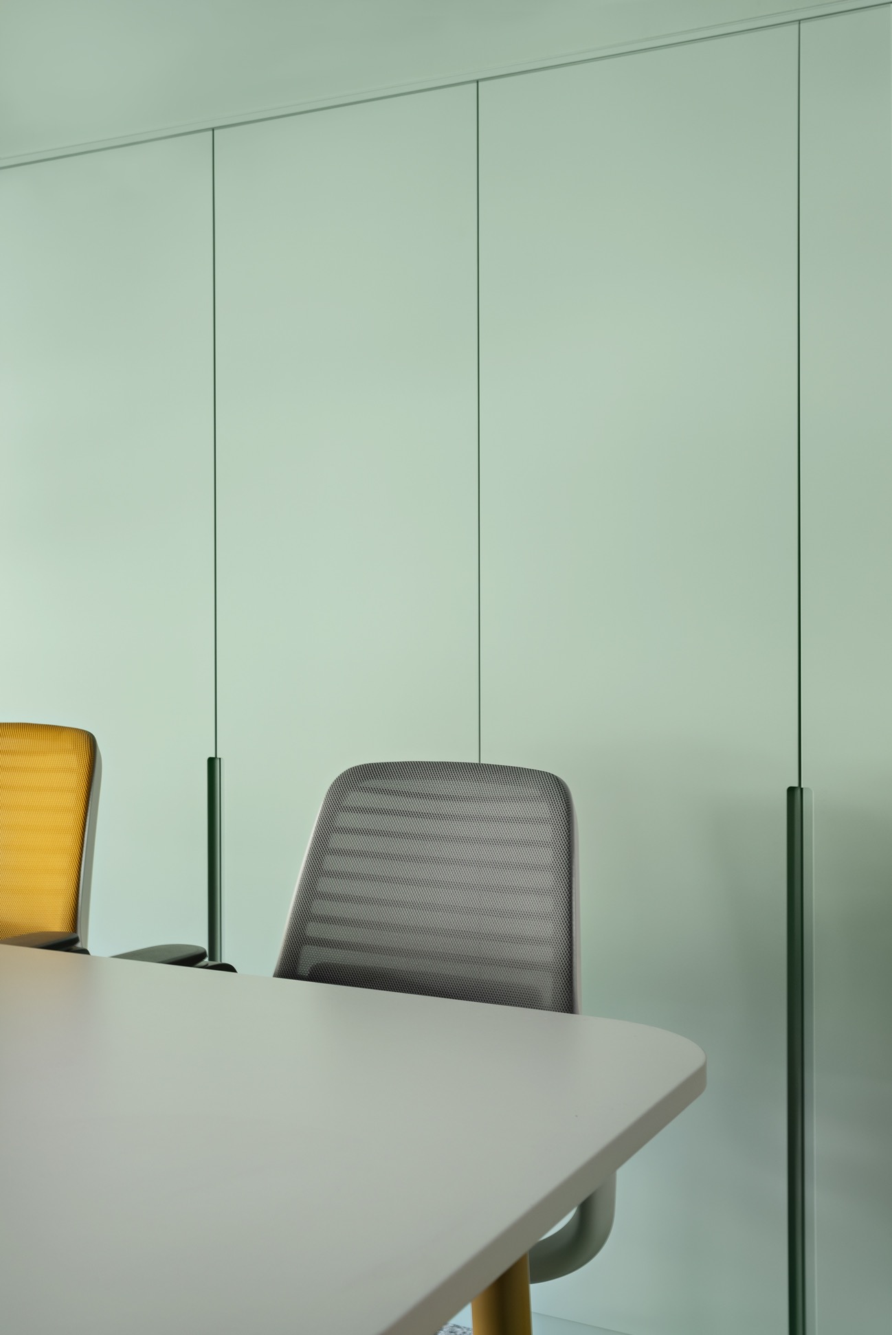 Minimalistyczne biuro: Jasnozielona ściana z ukrytymi szafami, szary stół z zaokrąglonymi rogami i krzesła z siatkowym oparciem w odcieniach szarości i żółci.