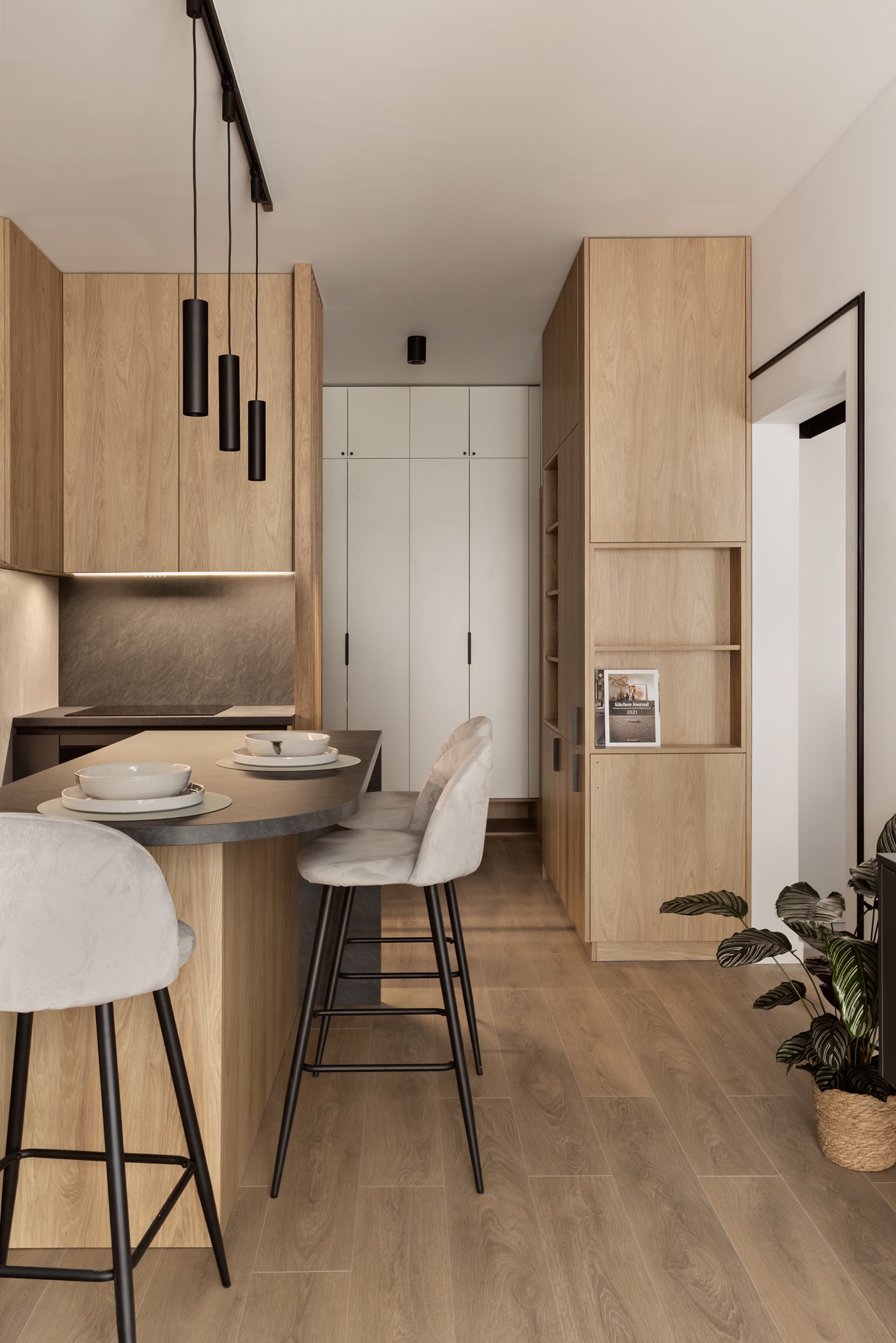 Nowoczesna kuchnia z drewnianymi szafkami, czarnym blatem i lampami. Dwa krzesła barowe z jasną tapicerką. Minimalistyczny design, ciepłe oświetlenie, roślina doniczkowa.