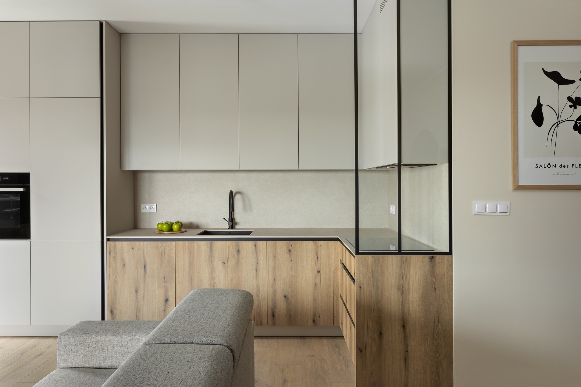 Nowoczesna kuchnia z drewnianymi frontami szafek dolnych i jasnymi górnymi, czarną armaturą i szklaną witryną. Na blacie zielone jabłka. Minimalistyczny plakat na ścianie.