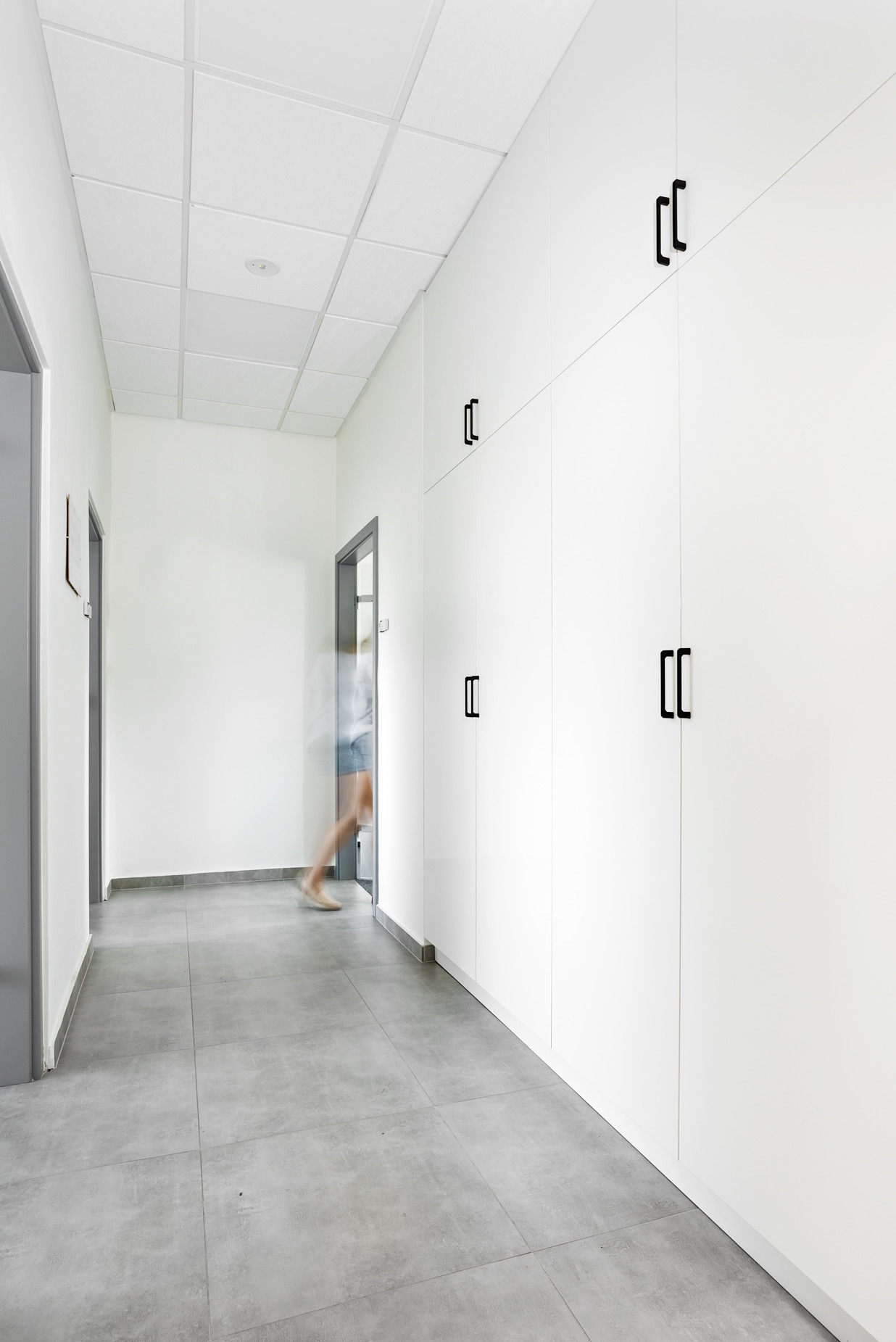 Białe szafy wnękowe z czarnymi uchwytami w korytarzu biurowym, widok z perspektywy podłogi, z rozmytą postacią w tle, szara podłoga z płytek.