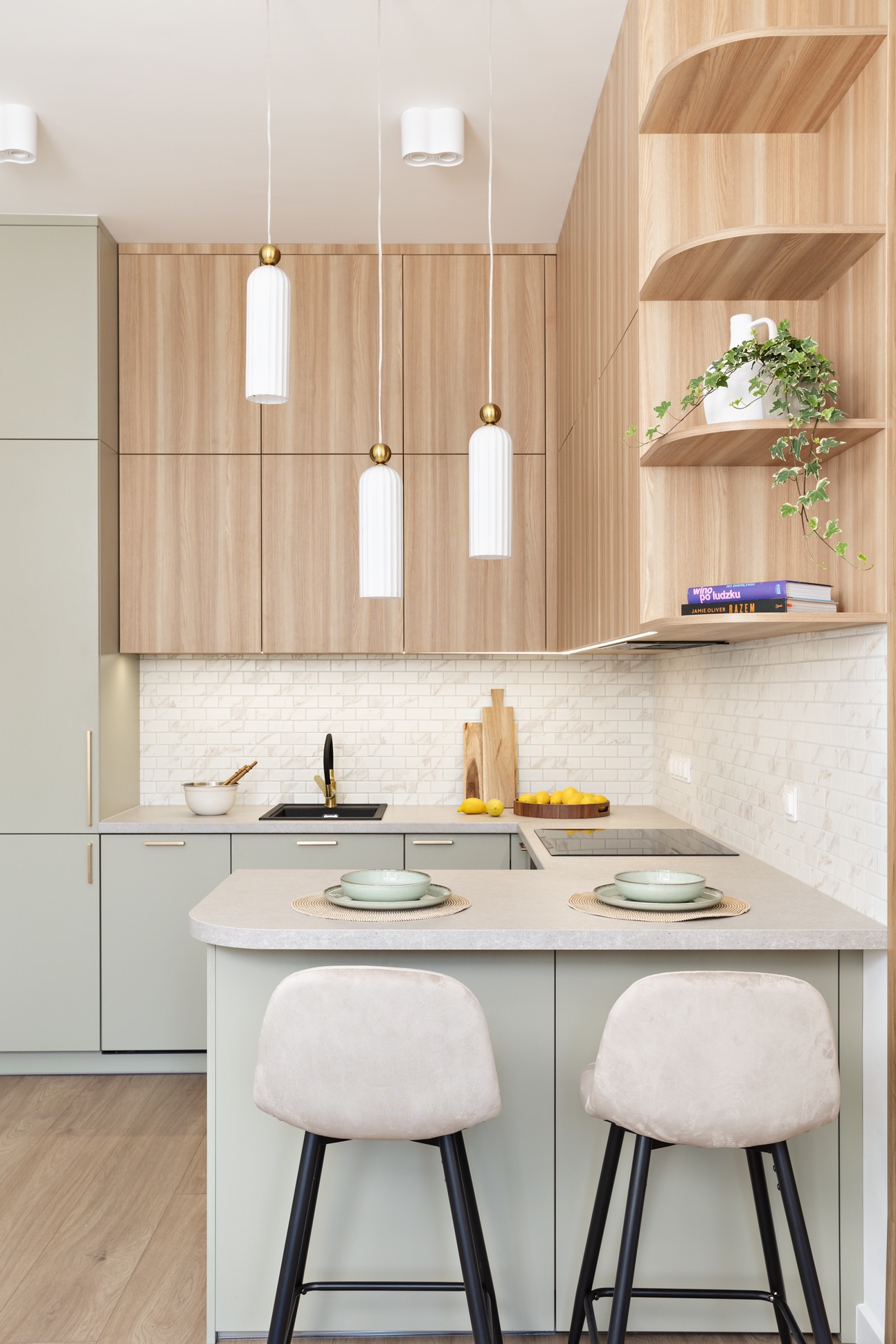 Jasna kuchnia z drewnianymi akcentami: szafki górne z forniru, otwarte półki z rośliną, minimalistyczne lampy wiszące i krzesła barowe z beżową tapicerką.