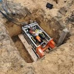 GROUND VISION SPÓŁKA Z OGRANICZONĄ ODPOWIEDZIALNOŚCIĄ - Maszyna do przewiertów w wykopie, widoczne kable i drewniane elementy. Ujęcie z góry, ziemisty kolor piasku.