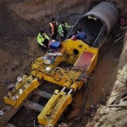 GROUND VISION SPÓŁKA Z OGRANICZONĄ ODPOWIEDZIALNOŚCIĄ - Żółta maszyna do przewiertów w wykopie, z widoczną rurą i grupą osób w odblaskowych kamizelkach, obserwujących proces. Ujęcie z góry, podkreślające głębokość wykopu.