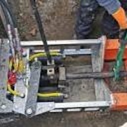 GROUND VISION SPÓŁKA Z OGRANICZONĄ ODPOWIEDZIALNOŚCIĄ - Przewiert sterowany: stalowa maszyna z przewodami w wykopie zabezpieczonym drewnem i pomarańczowymi elementami. Widoczne żółte rękawice ochronne.