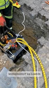 Praca przy wykopie: operator w rękawicach obsługuje maszynę do przewiertów sterowanych z żółtymi przewodami. Widoczne krawędzie wykopu i kostka brukowa.