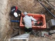 Pracownik w wykopie, w pomarańczowej skrzyni, wykonuje prace przy rurze. Widoczny fragment drabiny, ściany wykopu z piasku i narzędzia.