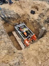 Maszyna do przewiertów w wykopie, widoczne kable i drewniane elementy. Ujęcie z góry, ziemisty kolor piasku.