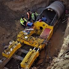 Żółta maszyna do przewiertów w wykopie, z widoczną rurą i grupą osób w odblaskowych kamizelkach, obserwujących proces. Ujęcie z góry, podkreślające głębokość wykopu.