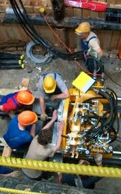 Praca przy maszynie do przewiertów sterowanych. Czterech robotników w kaskach obsługuje urządzenie. Widoczne kable i rury. Żółta maszyna z licznymi przewodami.