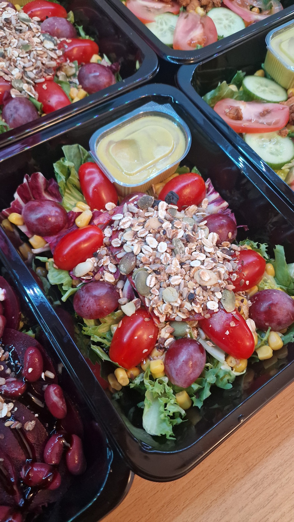 Sałatka z pomidorkami cherry, winogronami i ziarnami w czarnym pojemniku cateringowym, z sosem w osobnym pojemniczku. Zdjęcie z góry, widoczne inne pojemniki z jedzeniem.