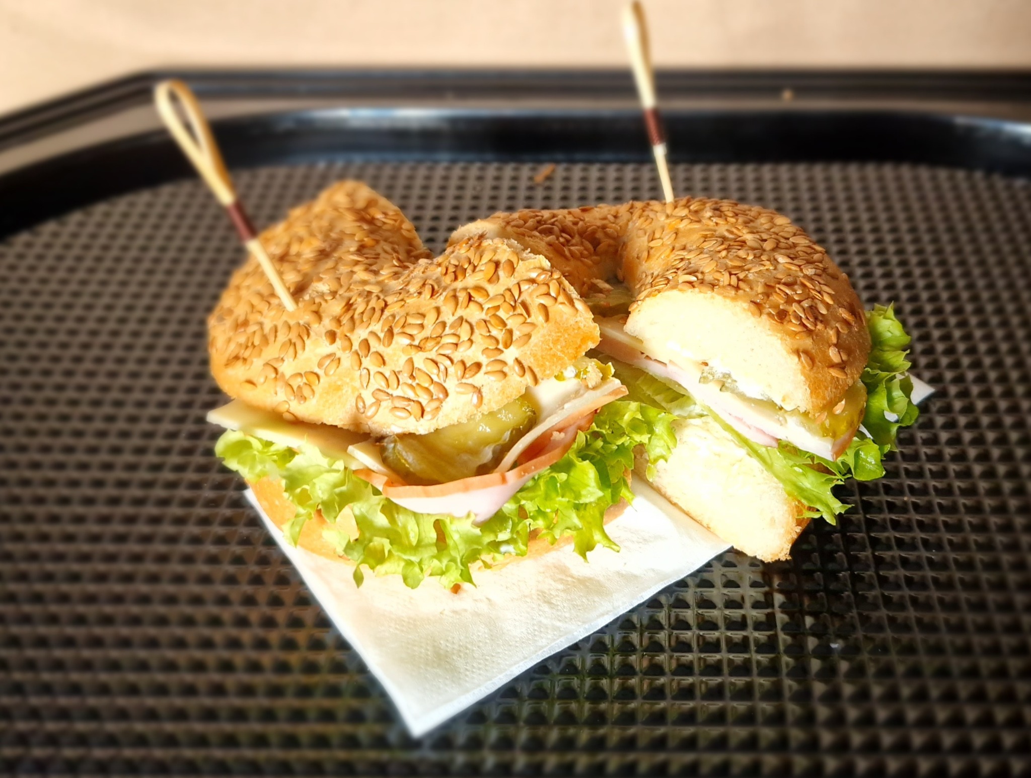 Przekrojony bajgiel z sezamem, szynką, serem, sałatą i ogórkiem konserwowym, spięty wykałaczkami, na serwetce i czarnej tacy. Idealny na catering.