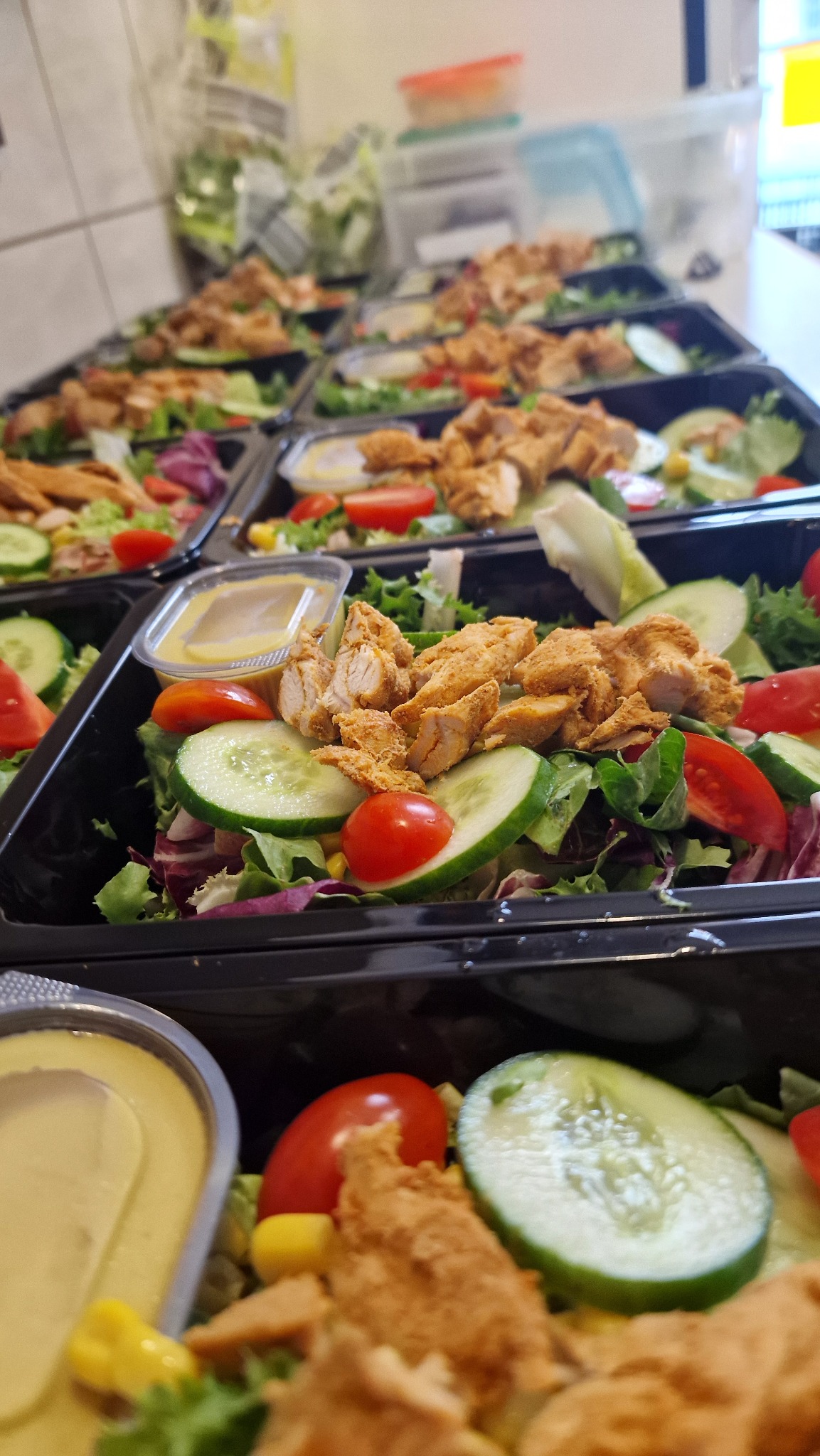 Rząd czarnych pojemników z sałatkami: zielone liście, pomidory, ogórki, kukurydza i kawałki kurczaka. Dodatkowo sos w małym pojemniku. Przygotowane porcje cateringowe.