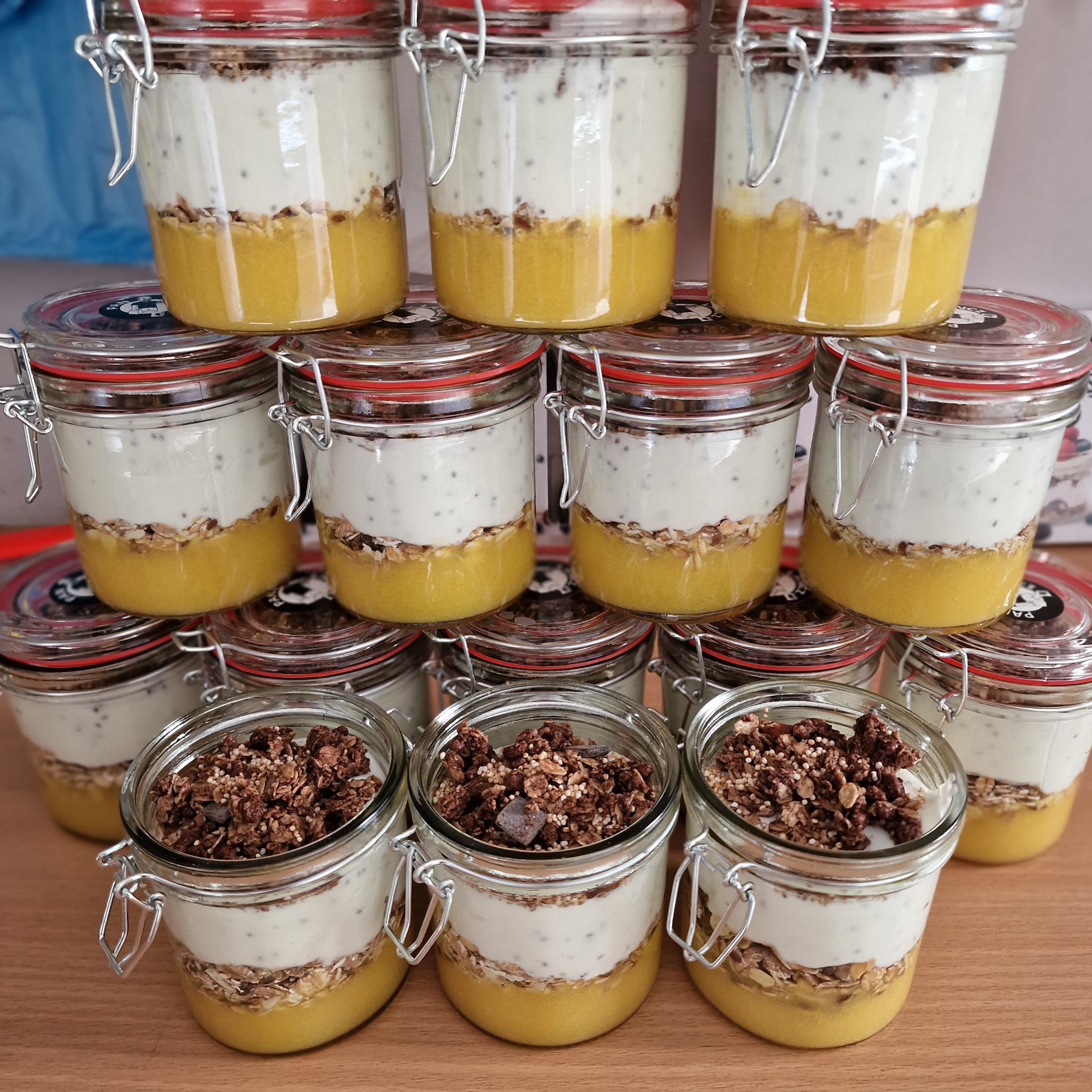 Deser w słoiczkach z warstwami: mus mango, granola, jogurt chia i kruszonka czekoladowa. Ułożone piramidalnie na drewnianym stole. Catering dietetyczny.