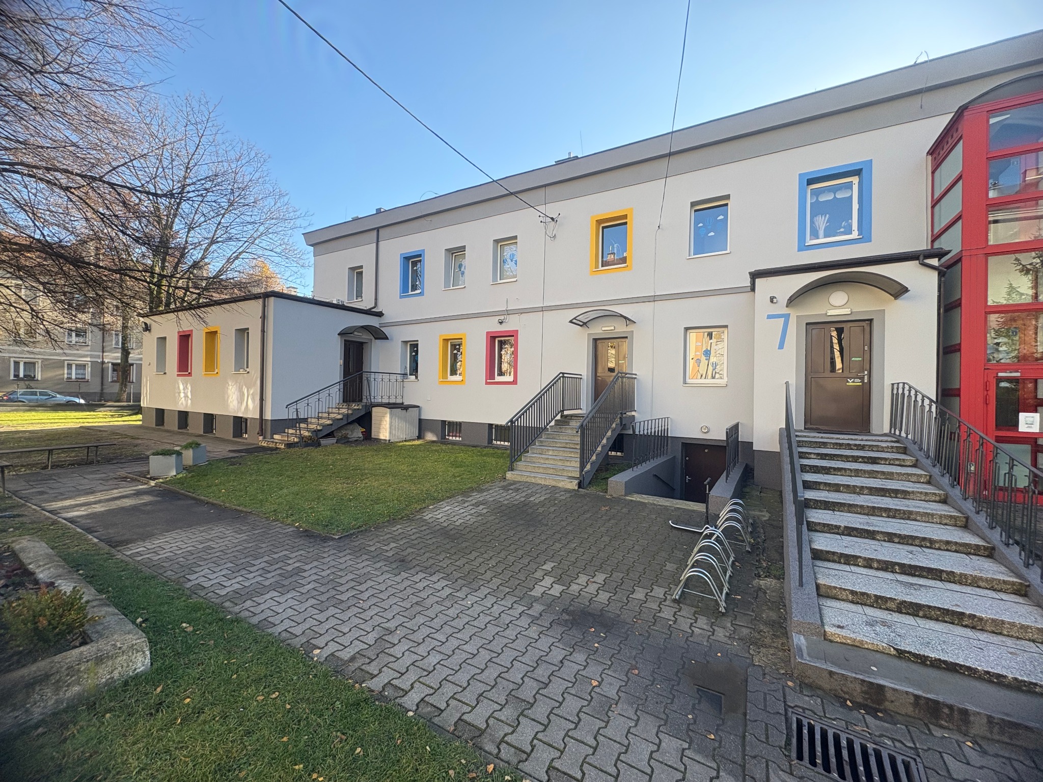 Dwupiętrowy budynek żłobka z kolorowymi ramami okiennymi i numerem 7 przy wejściu, otoczony chodnikiem z kostki i zielenią. Widok na fasadę w słoneczny dzień.