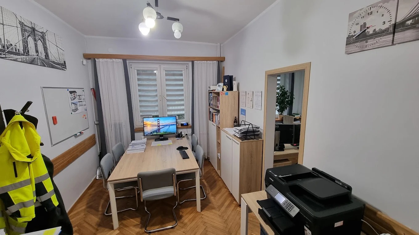 Biuro projektowe: stół z komputerem i planami, krzesła, regał z dokumentami, drukarka. Na ścianie tablica i grafika z mostem. Żółta kurtka robocza na wieszaku.