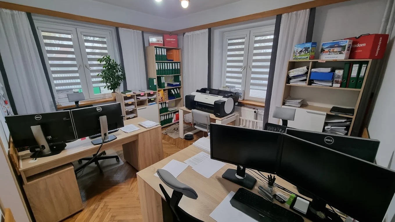 Biuro projektowe z dwoma stanowiskami pracy, monitorami Dell, drukarką Canon i regałami z segregatorami. Widok na okna z żaluzjami i rośliną doniczkową.