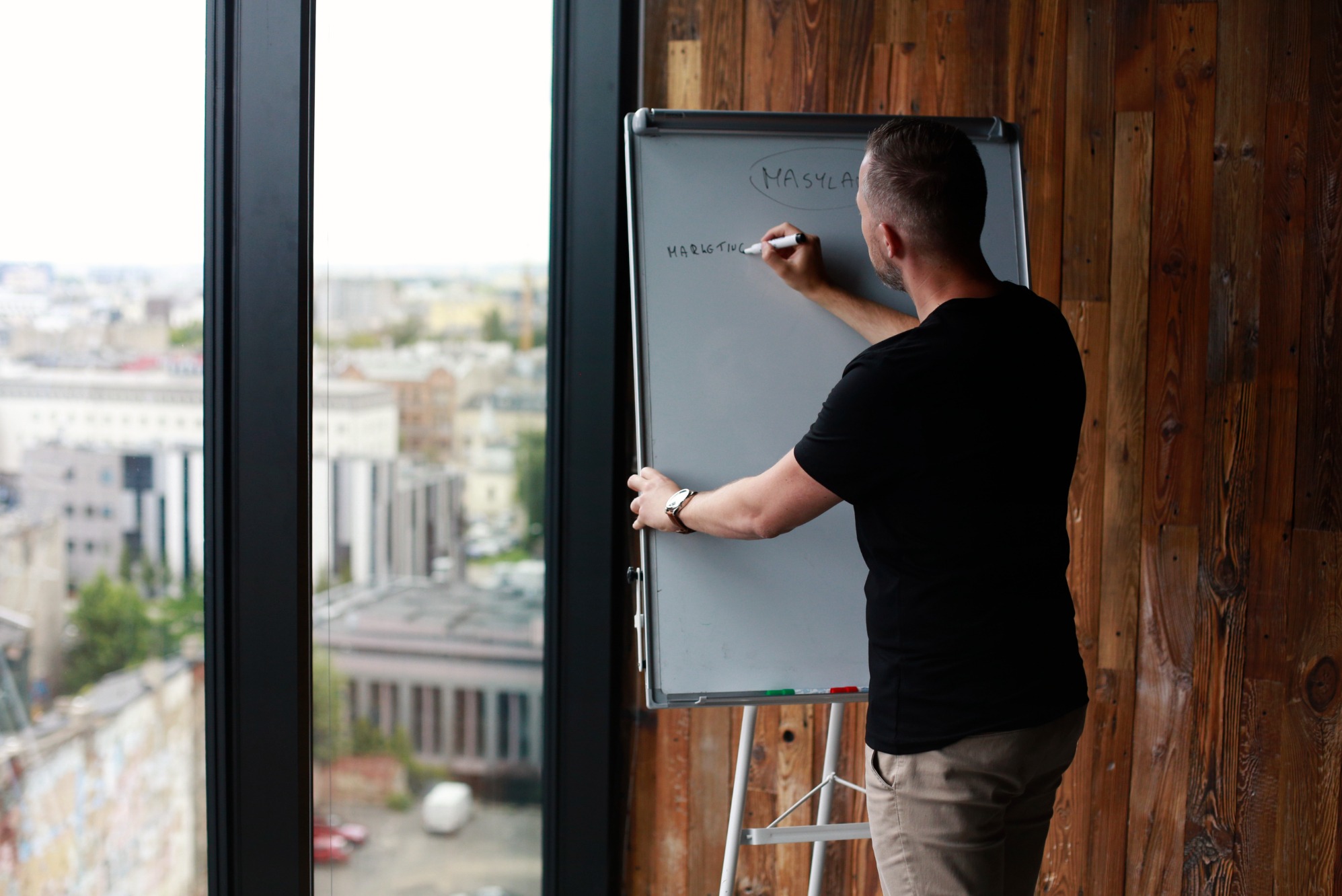 Mężczyzna pisze markerem na flipcharcie słowo 'Marketing' w jasnym biurze z widokiem na miasto. Prezentacja strategii marketingowej na tle drewnianej ściany.