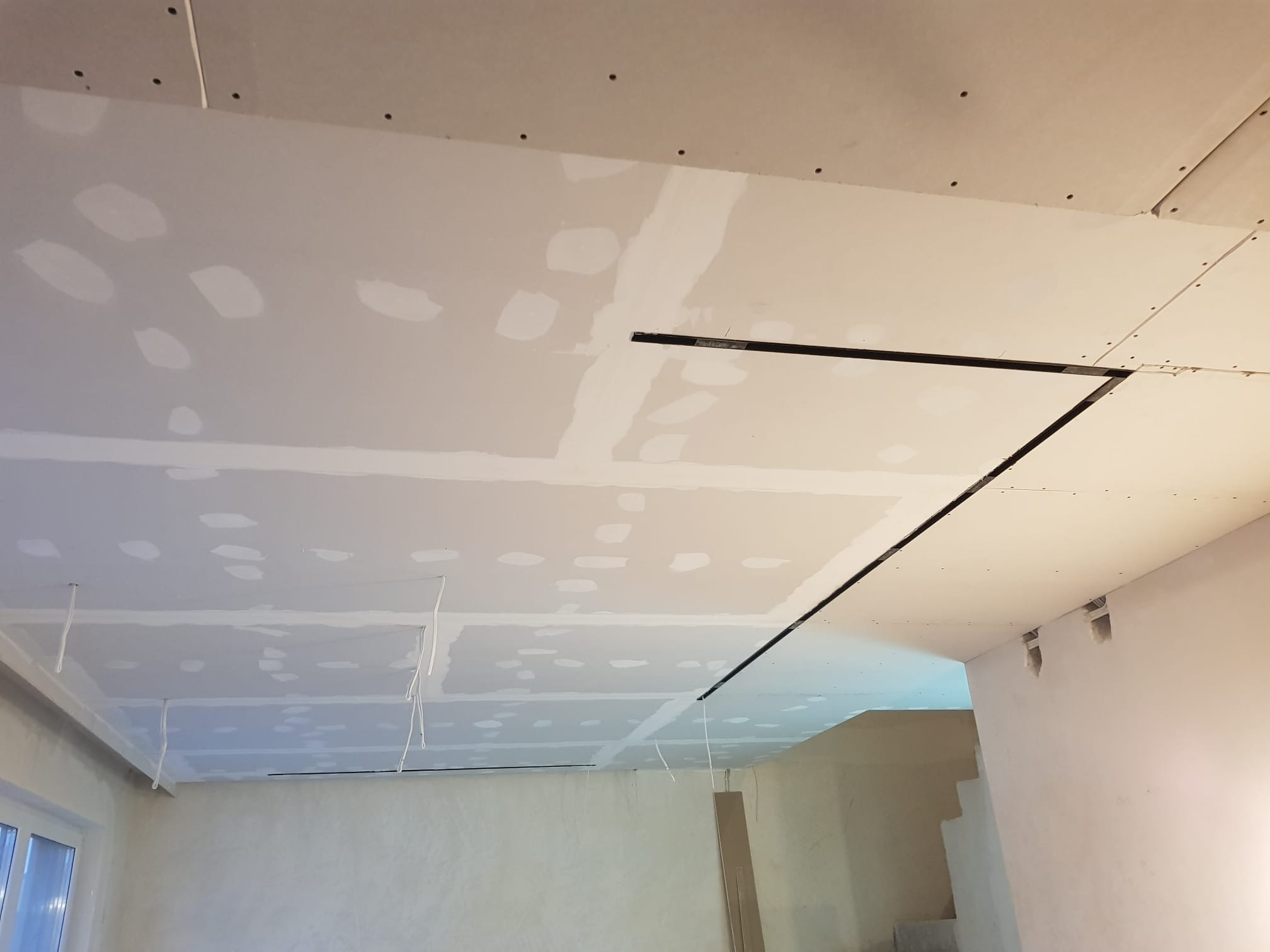 Sufit podwieszany z płyt gipsowo-kartonowych w trakcie szpachlowania, widoczne łączenia i otwory na oświetlenie. Prace wykończeniowe w jasnym pomieszczeniu.