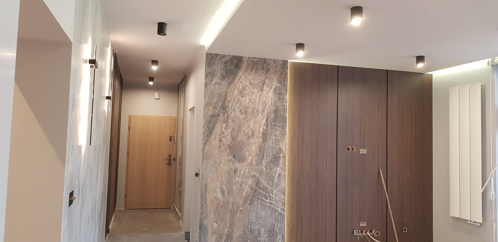 Nowoczesny korytarz po remoncie: marmurowa ściana, drewniana zabudowa, punktowe oświetlenie sufitowe i ścienne. Widoczne gniazdka elektryczne i grzejnik.