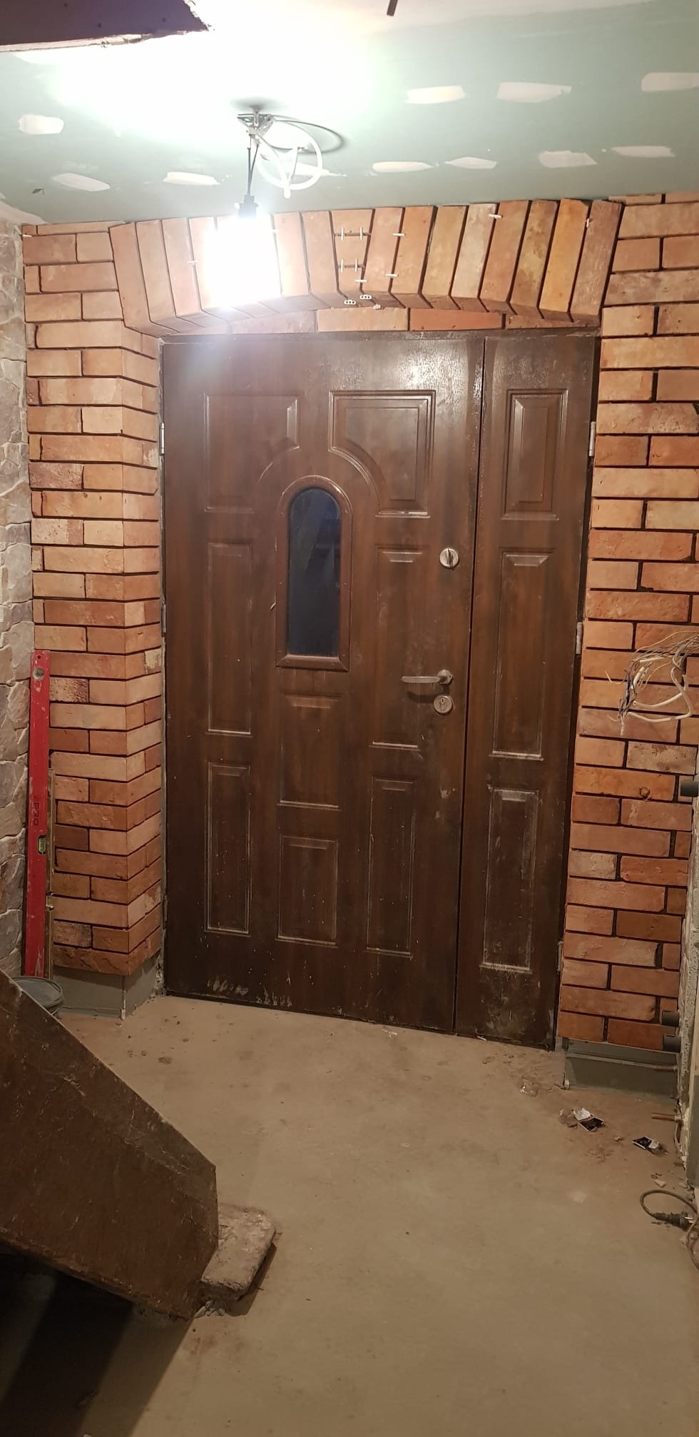 Ciemne, drewniane drzwi wejściowe z małym okienkiem, otoczone ceglanym łukiem i ścianą z cegły klinkierowej. Widoczne ślady prac wykończeniowych i surowy stan podłogi.