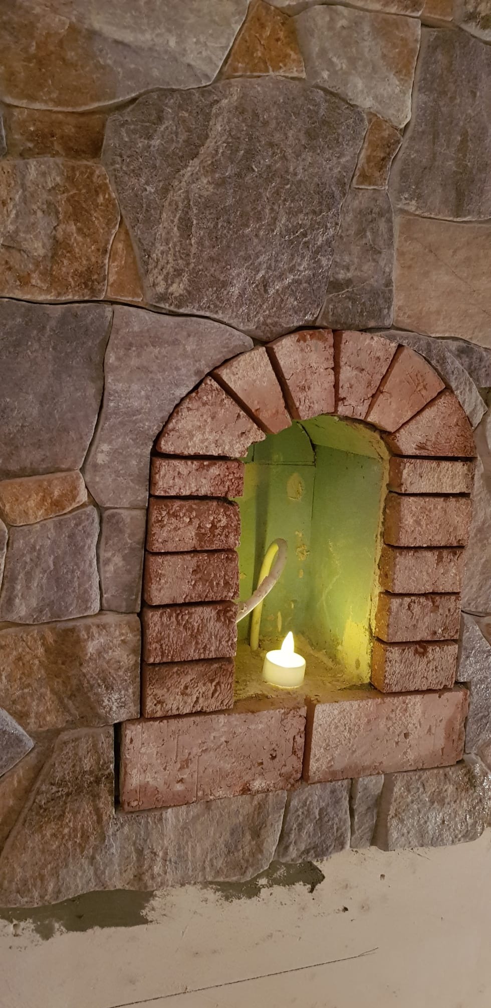 Kamienna ściana z ceglaną wnęką w kształcie łuku, w której umieszczona jest zapalona świeczka. W tle widoczna zielona ściana wewnątrz wnęki.