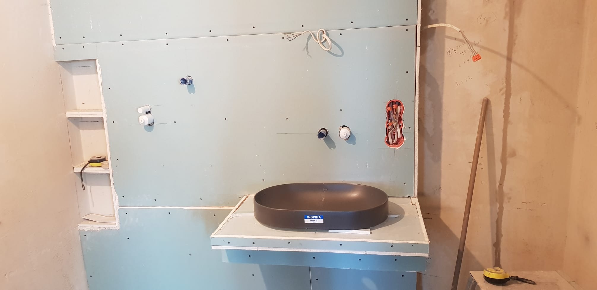 Montaż umywalki Inspira Roca na tle płyty gipsowej z instalacją wodną i elektryczną. Widoczny etap prac wykończeniowych w łazience.