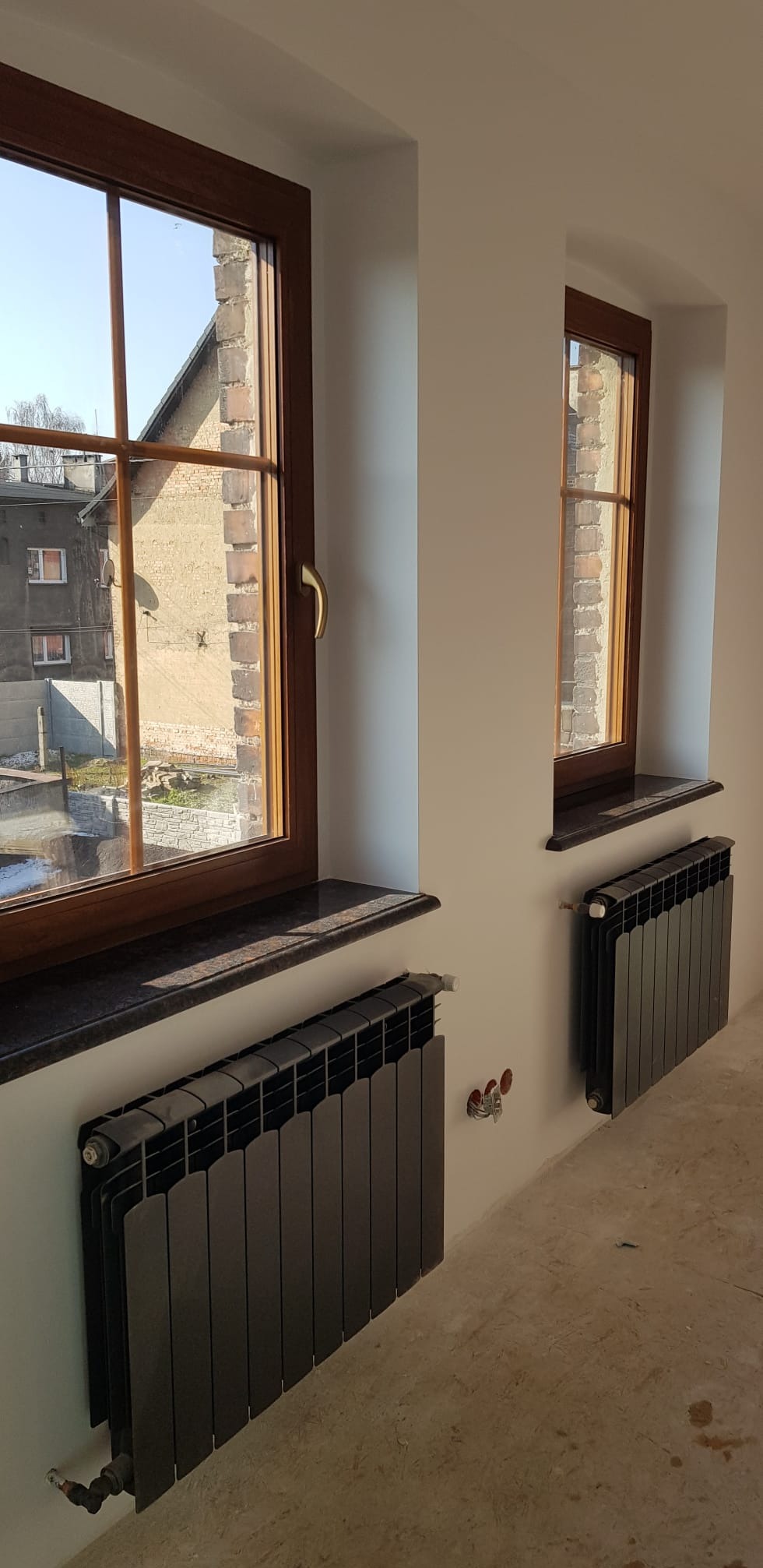 Dwa okna z drewnianą ramą i czarnym parapetem, pod nimi grzejniki w kolorze grafitowym. Białe ściany i surowa podłoga. Widok na budynek z cegły za oknem.