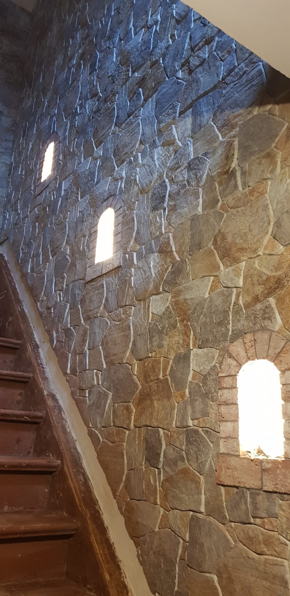 Kamienna ściana z trzema oknami w kształcie łuków, oświetlonych od wewnątrz. Obok widoczne drewniane schody. Dekoracyjne wykończenie wnętrza.