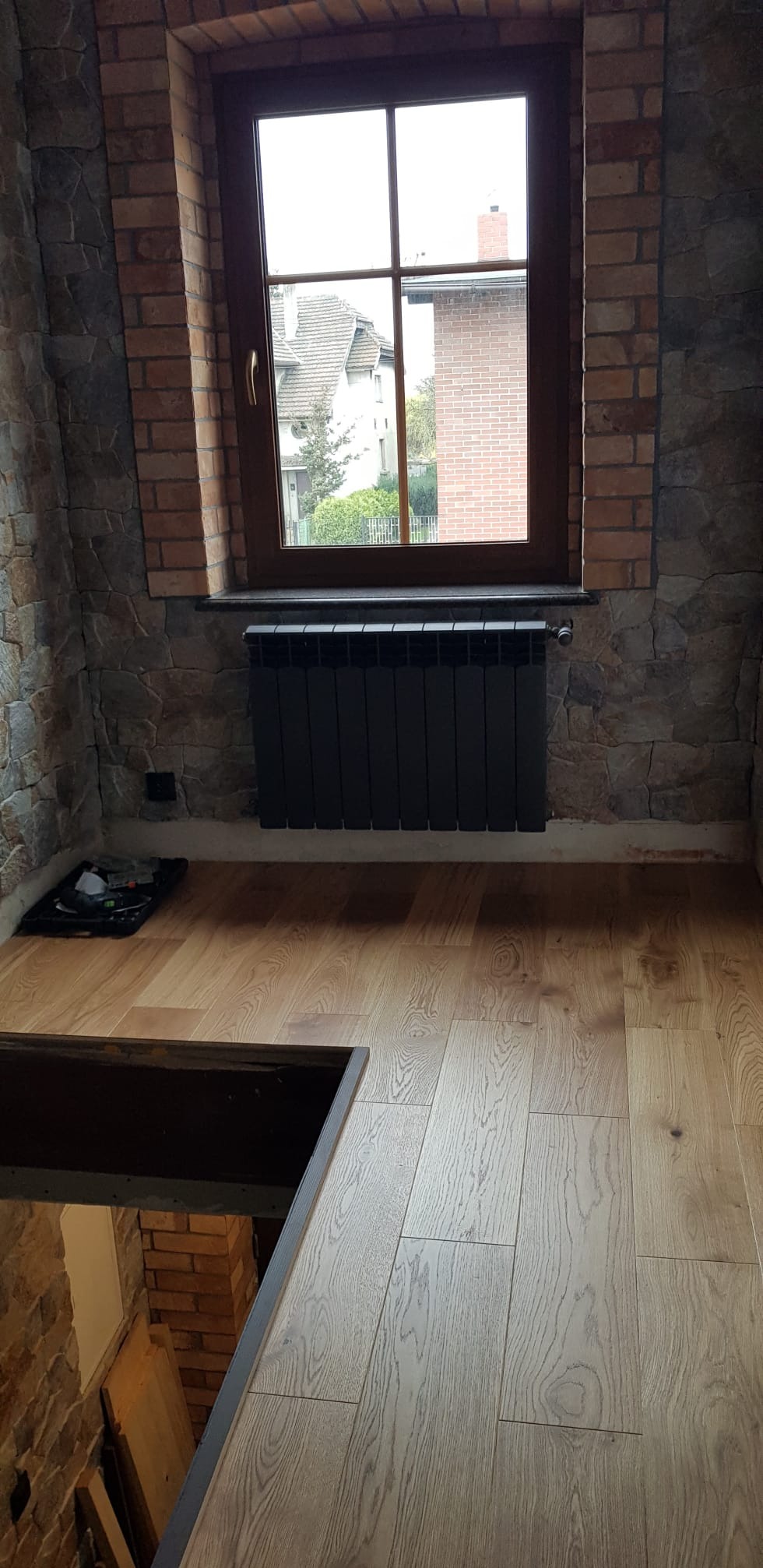 Pokój z oknem, grzejnikiem i panelami podłogowymi. Ściany wykończone cegłą i kamieniem. Widoczny fragment schodów. Ujęcie z góry, podkreślające fakturę podłogi.