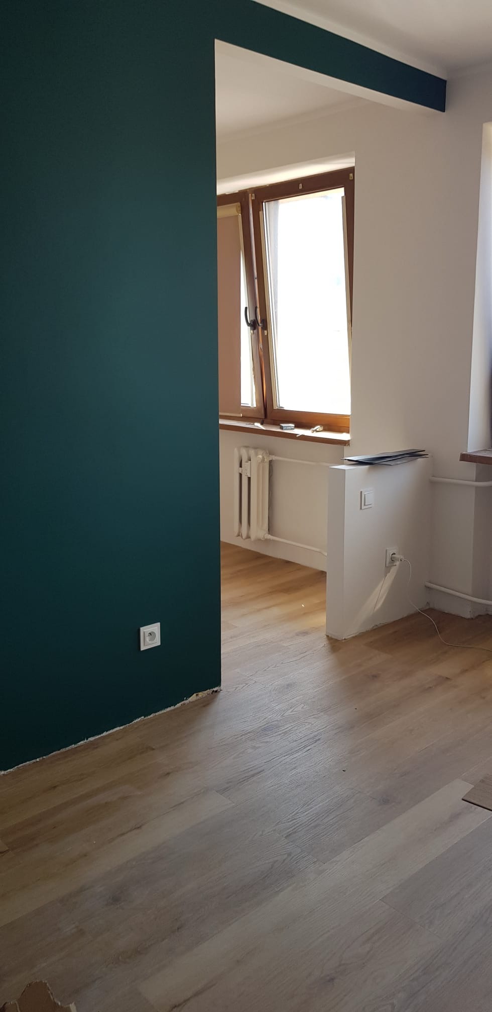 Świeżo pomalowane wnętrze z ciemnozieloną ścianą i jasną podłogą. Widoczne okno z grzejnikiem i fragment białej ściany. Gniazdko elektryczne na ciemnej ścianie.