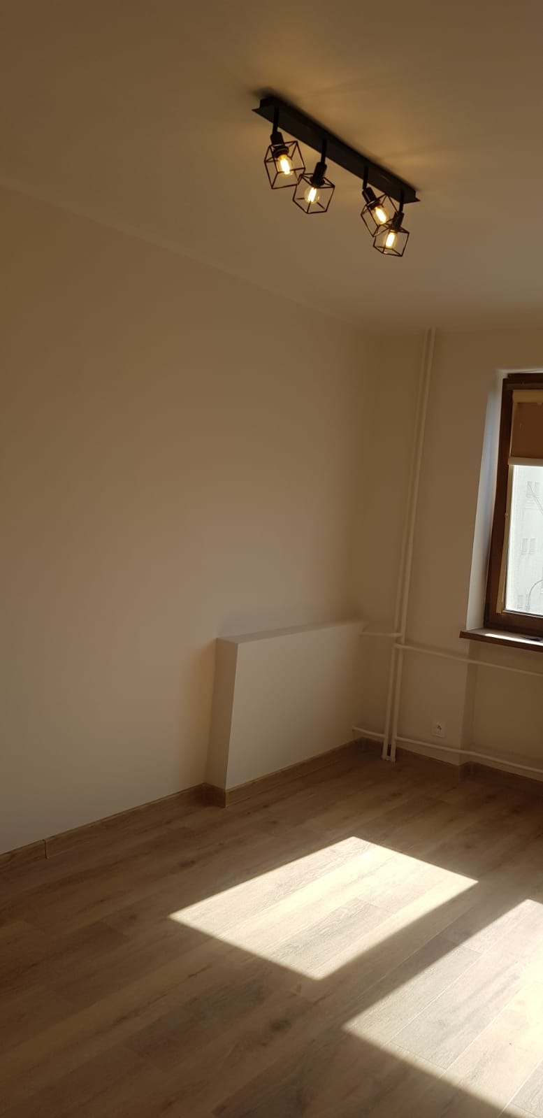 Jasne wnętrze po remoncie: drewniana podłoga, świeżo pomalowane ściany w odcieniach beżu, nowoczesna lampa sufitowa z geometrycznymi kloszami i okno z drewnianą ramą.