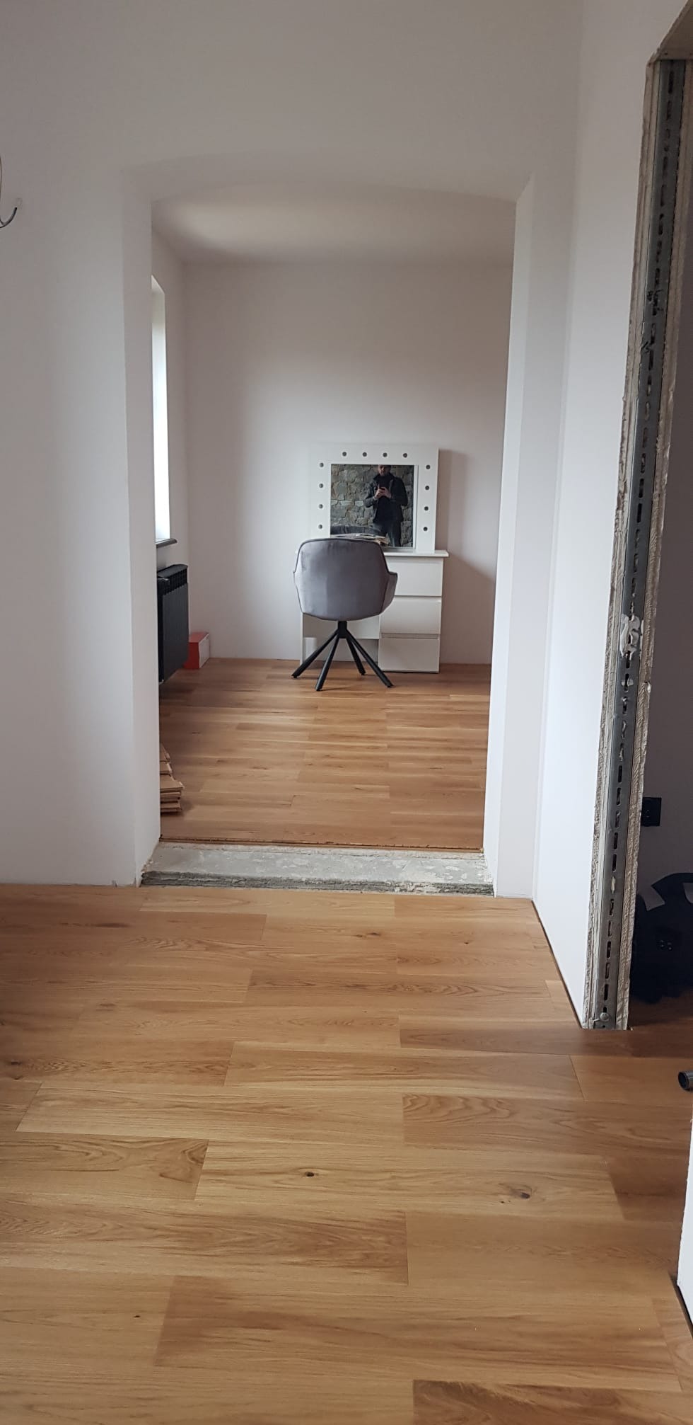 Jasne panele podłogowe w nowym wnętrzu, widok z korytarza na pokój z toaletką i krzesłem. Fragment metalowej ościeżnicy po prawej stronie kadru.