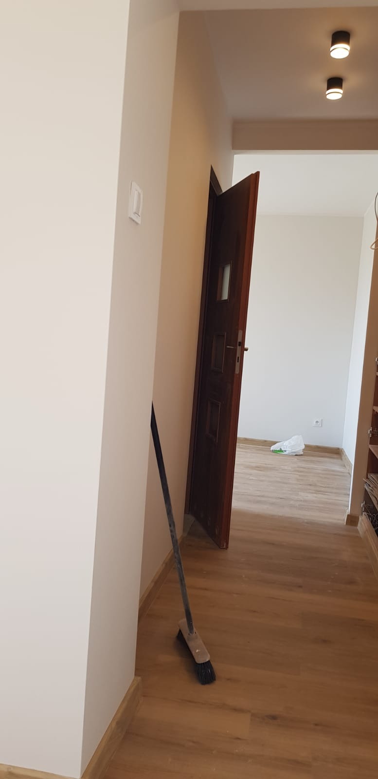 Jasne wnętrze po odświeżeniu: pomalowane ściany, drewnopodobna podłoga, otwarte drzwi. W rogu stoi miotła, a na suficie widoczne nowoczesne lampy.