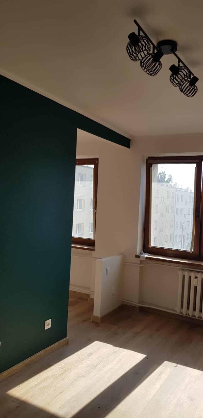 Pomieszczenie po malowaniu: ściana w odcieniu butelkowej zieleni kontrastuje z bielą, drewniane okna, parkiet i nowoczesna lampa sufitowa.