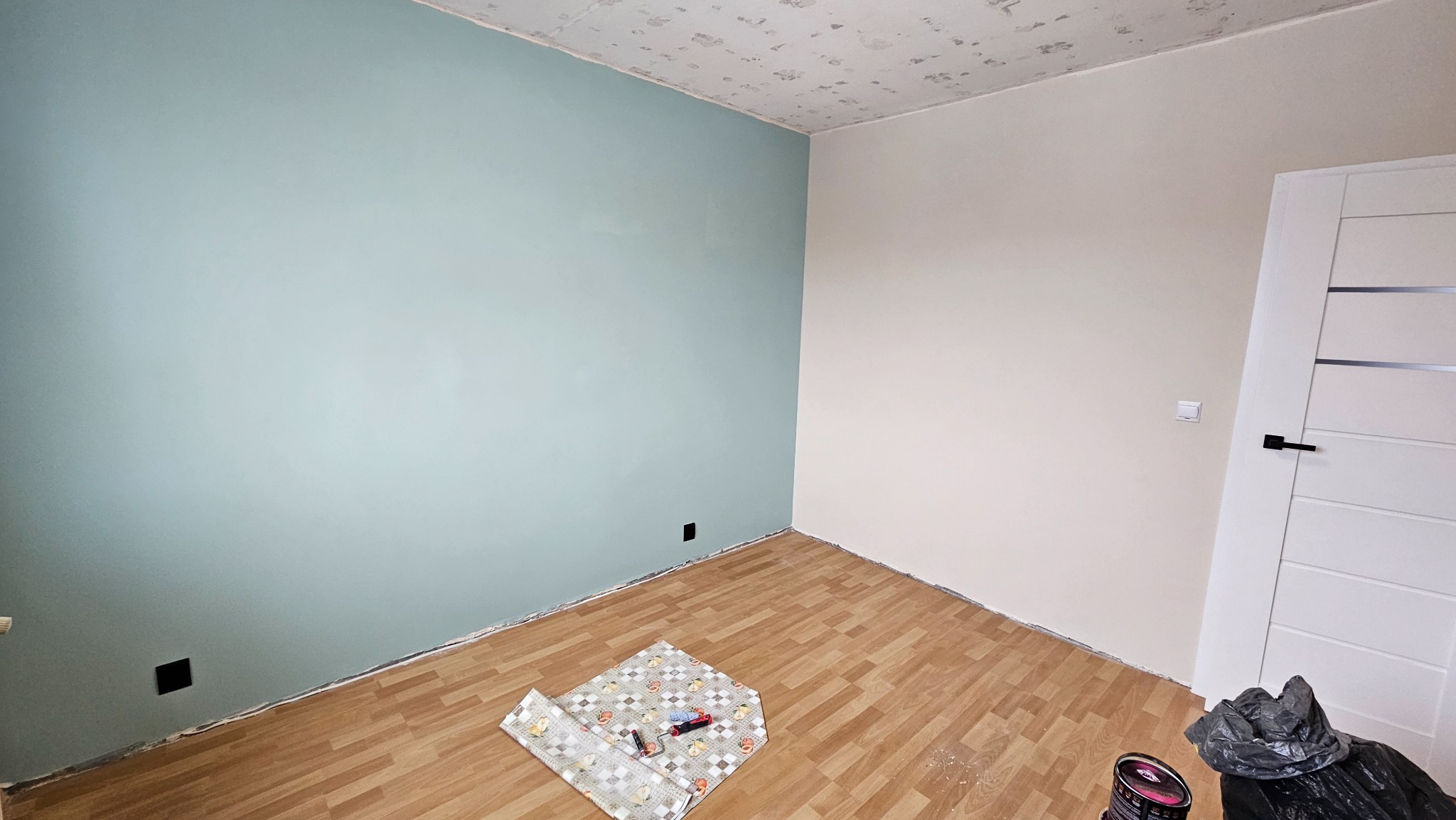 Pokój po malowaniu: ściana w odcieniu błękitu i beżu, drewniana podłoga. Widoczne narzędzia malarskie i puszka farby. Sufit z widocznymi śladami po remoncie.