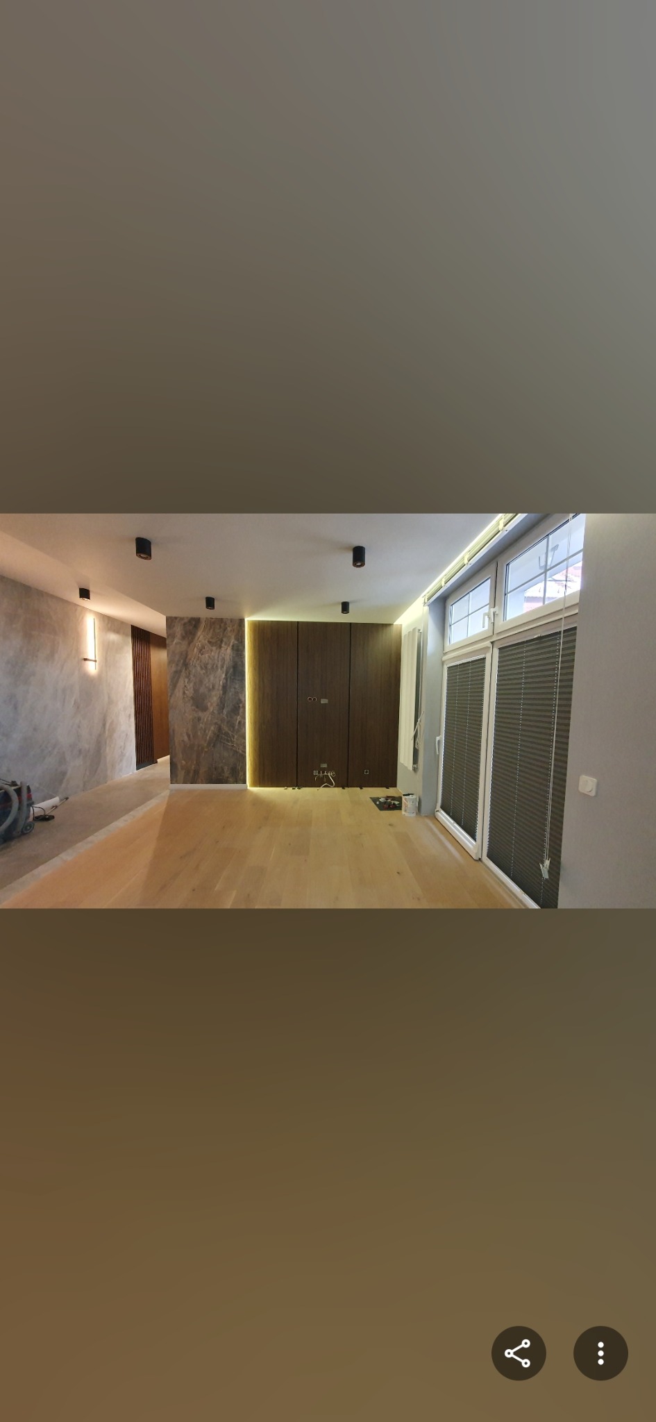 Nowoczesne wnętrze z drewnianą podłogą, szarą ścianą z efektem betonu i zabudową meblową z podświetleniem LED. Widoczne okna z żaluzjami.