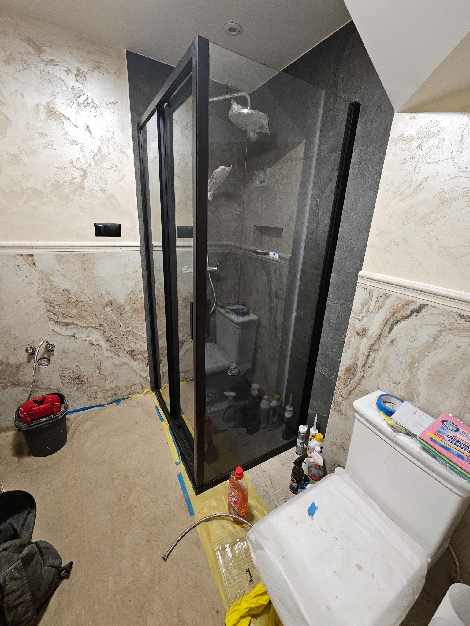 Nowoczesna kabina prysznicowa z czarnymi profilami w łazience z marmurowymi ścianami i zabezpieczonymi elementami podczas prac wykończeniowych.