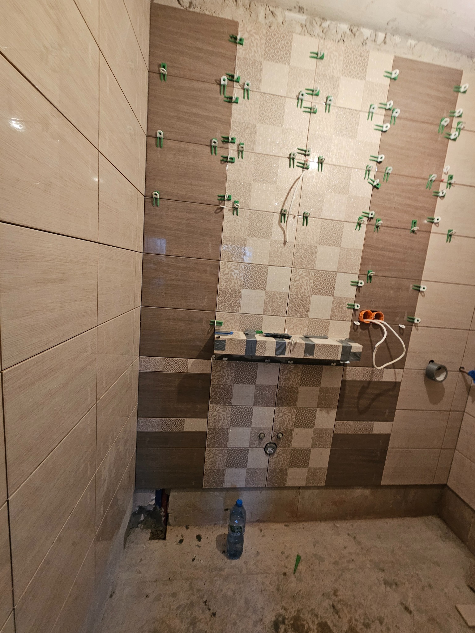 Układanie płytek w łazience z użyciem klipsów poziomujących. Widoczne różne wzory płytek w odcieniach beżu i brązu. Częściowo wykończona ściana, surowy stan podłogi.