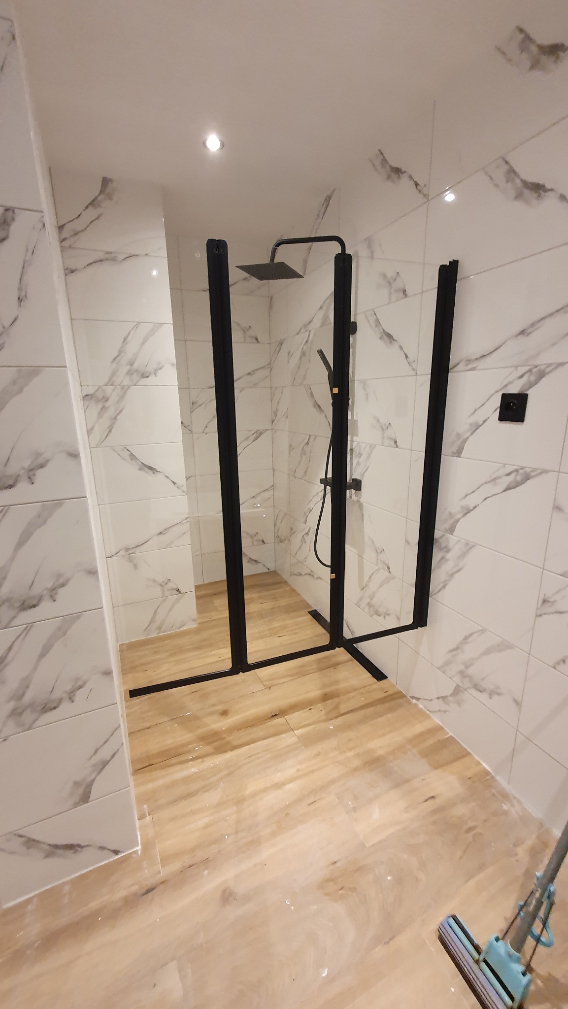 Nowoczesna kabina prysznicowa z czarnymi profilami i deszczownicą, wykończona płytkami imitującymi marmur. Podłoga wyłożona panelami imitującymi drewno.